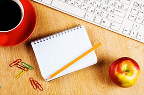 A cup of coffee, a keyboard, paper clips, a notebook, a pencil and an apple sit on a desk